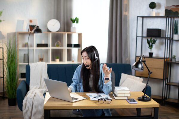 Chinesische Frau mit Headset, die während eines Videochats spricht und gestikuliert, während sie einen Laptop für Fernarbeit benutzt
