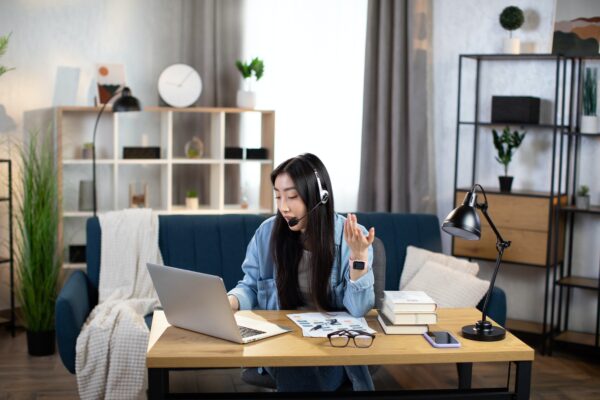 Chinese woman in headset talking and gesturing during video chat, using laptop pc for distance work