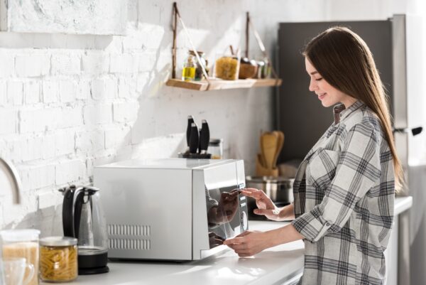 vista lateral de una mujer sonriente con camisa usando el microondas en la cocina